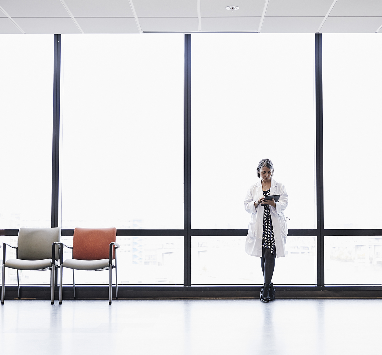 A woman in a lab coat leans again a large office while on a tablet in an empty waiting room.